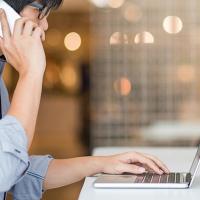 A man sits at a table while talking on his cell phone and typing on his laptop.
